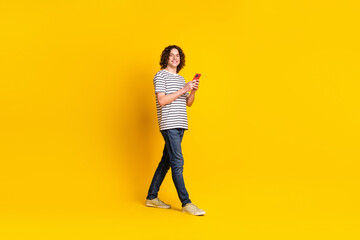 Fototapeta premium Full body portrait of nice young man walk empty space use phone wear striped t-shirt isolated on yellow color background