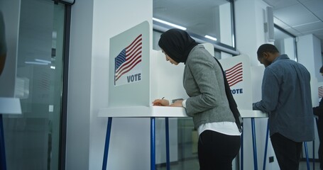 Muslim woman in hijab comes to vote in booth in polling station office. National Election Day in the United States. Political races of US presidential candidates. Concept of civic duty. Dolly shot. - obrazy, fototapety, plakaty