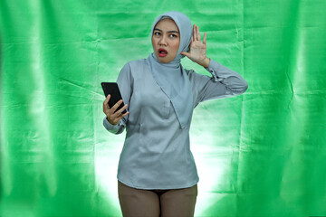 serious looking asian woman wearing hijab trying to overhear secret conversation and holding smartphone over green background
