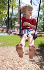 barefoot boy 6-7 years old sits on a sports net in a park on a playground on a summer day. children's playground in the city park, entertainment and recreation for children. Focus on children's feet