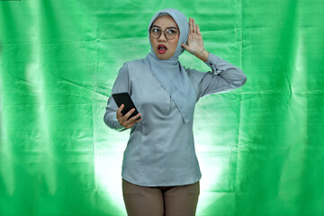 serious looking asian woman wearing hijab, glasses and blouse trying to overhear secret conversation and holding smartphone over green background
