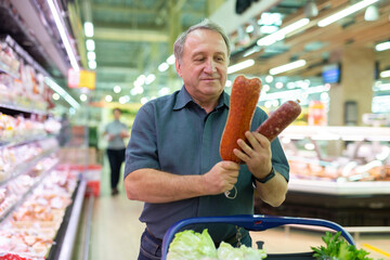 Mature man diligently choosing tasty sausage in supermarket