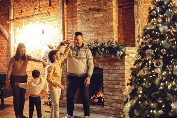 Happy family parents with two kids dancing together near fireplace and xmas tree in cozy...