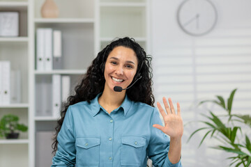 A cheerful woman in a blue shirt and headset waves to the camera, portraying a welcoming online customer service environment in a modern office. - obrazy, fototapety, plakaty