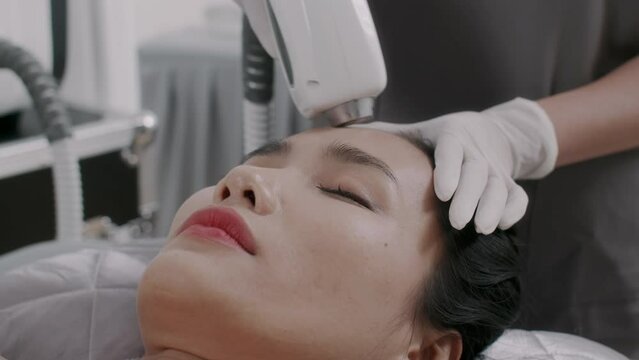 Close up of Asian woman getting rejuvenation laser treatment on forehead while visiting beauticians room