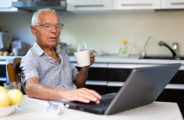 Handsome old man in glasses with mug of tea uses laptop while sitting on chair