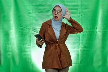 serious looking asian woman wearing hijab, glasses and blazer trying to overhear secret conversation and holding smartphone over green background

