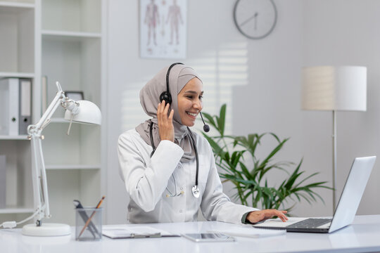 Cheerful female doctor in hijab sitting at her desk in a modern clinic, providing consultation via headset and laptop, with a warm smile.