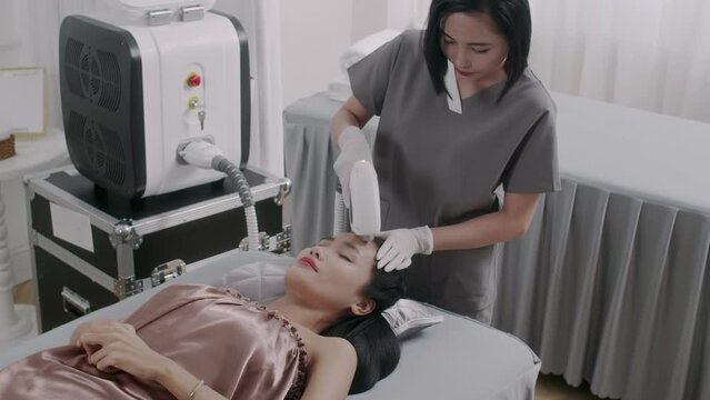 Medium shot of Asian beauty therapist giving laser treatment on face of young female client in clinic