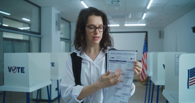 American woman speaks on camera, shows paper bulletin, calls for voting. National Elections Day in the United States. Voting booths at polling station. Concept of civic duty and patriotism. Portrait.