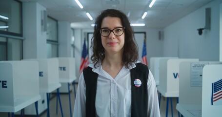 Obraz premium American female voter or polling officer speaks on camera, calls for voting. National Elections Day in the United States. Voting booths in the background at polling station. Civic duty and patriotism.