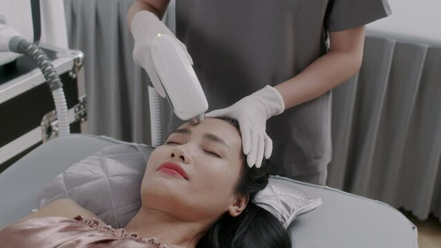 Medium close up of young woman of Asian ethnicity having anti-wrinkle laser treatment on forehead visiting beauty salon