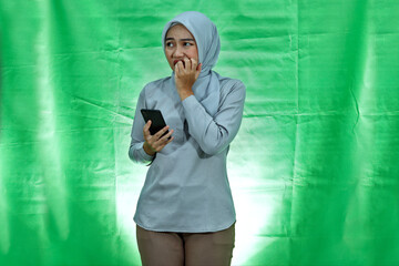 Young Asian woman wearing hijab and blouse holding smartphone looks anxious and worried afraid of something isolated over green background
