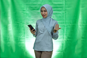 cheerful asian woman wearing hijab and blouse taunting at front and holding smartphone over green background
