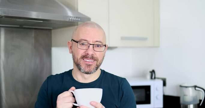 Footage of a middle aged man with a shaven head and a scruffy beard wearing glasses and a blue t-shirt drinking a cup of coffee or tea at home in a kitchen