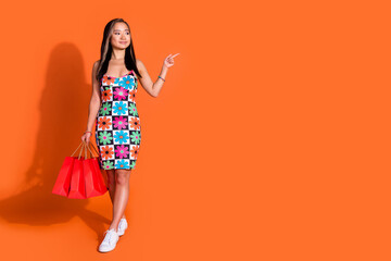 Full length photo of korean girl dressed flower print clothes look directing empty space hold bags isolated on orange color background