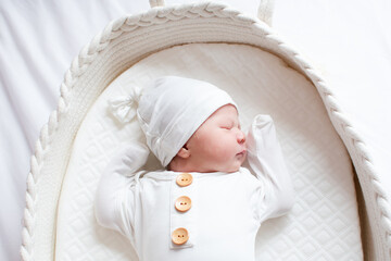 Newborn sleeping in a Moses basket wearing white pajamas and white hat. Sleeping newborn baby girl
