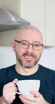 Portrait footage of a middle aged man with a shaven head and a scruffy beard wearing glasses and a blue t-shirt drinking a cup of coffee or tea at home in a kitchen