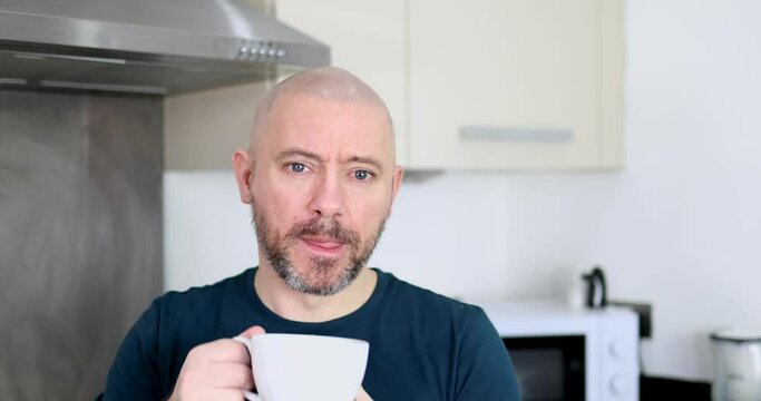 Footage of a middle aged man with a shaven head and a scruffy beard wearing a blue t-shirt drinking a cup of coffee or tea at home in a kitchen