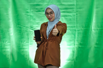 cheerful young Asian woman wearing hijab, glasses and blazer using a mobile phone and gesturing thumbs up isolated on white background
