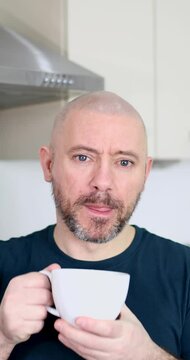 Portrait footage of a middle aged man with a shaven head and a scruffy beard wearing a blue t-shirt drinking a cup of coffee or tea at home in a kitchen