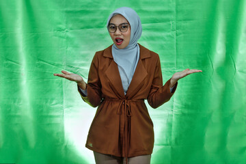 smiling young Asian woman wearing hijab, glasses and blazer showing copy space with both palms isolated on green background
