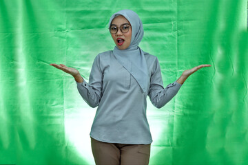 smiling young Asian woman wearing hijab, glasses and blouse showing copy space with both palms isolated on green background
