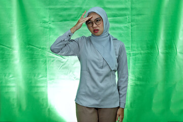 young Asian woman wearing hijab, glasses and blouse looking dizzy and having headache on green background
