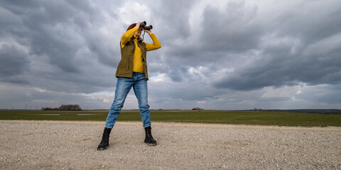 female ornithologist birdman or explorer watches birds with binoculars against a background of a stormy sky - 788369674