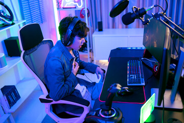 Young Asian man professional gamer wearing a jeans jacket sits on a chair and grabs his chest suffering heart attack from stress playing a game or competition. Concept of pain from gaming and esports.