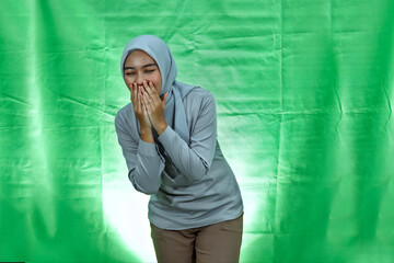 laughing young Asian woman wearing hijab and blouse covering mouth with hand on green background
