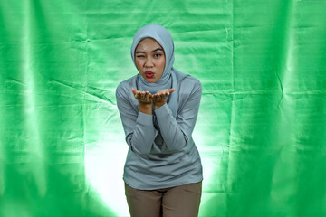 charming young Asian woman wearing hijab and blouse hold hand palms send air kiss to boyfriend over green background

