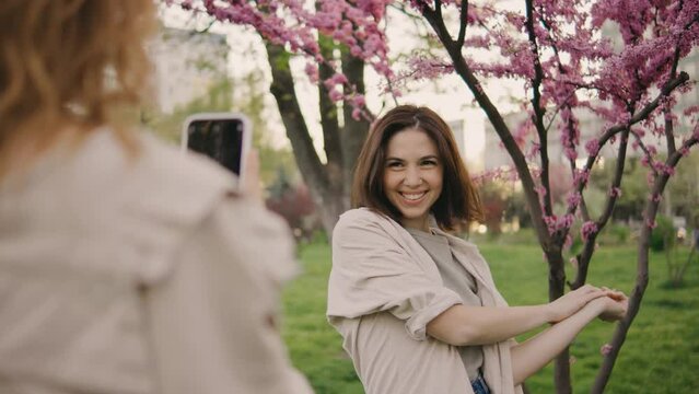 two beautiful young women taking pictures of each other on a smartphone against the background of pink blossoming cherry or sakura trees. Girls laugh, rejoice and make faces at the camera