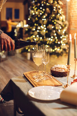 New Years Eve celebration. Man pouring champagne into glasses standing on table with festive xmas dinner, candles and wrapped gifts against blurred background of brick fireplace and firtree, cropped
