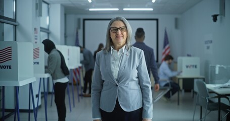 Businesswoman stands in a modern polling station, poses, looks at camera, smiles. Portrait of a mature woman, United States of America elections voter. Background with voting booths and American flag.