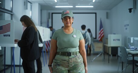 Woman in camouflage uniform stands in polling station and looks at camera. Portrait of female soldier, United States of America elections voter. Background with voting booths. Concept of civic duty.