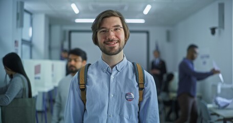 Portrait of Caucasian man, United States of America elections voter. Man with badge stands in modern polling station, poses, smiles, looks at camera. Background with voting booths. Civic duty concept. - obrazy, fototapety, plakaty