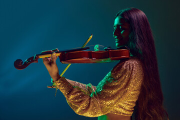 Cheerful young Indian woman, violinist in festive attire performing in neon light against gradient...