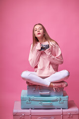 Travel concept. Studio portrait of pretty young woman holding camera sitting on baggage valises. Pink background.
