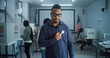 Portrait of African American man, United States of America elections voter. Man with badge stands in modern polling station, poses, looks at camera. Background with voting booths. Election Day in USA. - obrazy, fototapety, plakaty