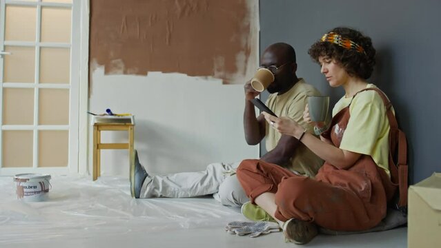 Side full footage of diverse married couple resting with cup of tea or coffee while sitting on floor in remodeled room, Caucasian wife showing funny pictures to Black husband