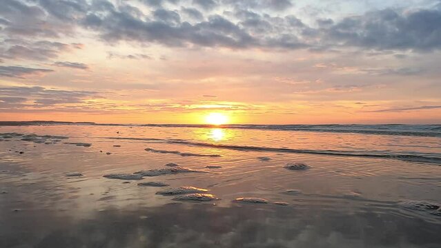 This slow-motion footage captures the tranquil beauty of a sunset on a serene beach, with gentle waves lapping against the shore. The golden sun dips toward the horizon, reflecting its warm hues on