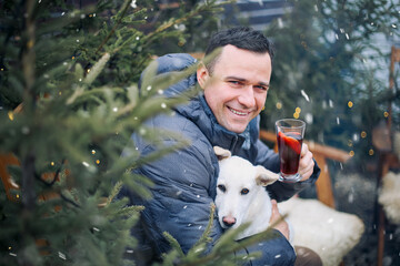 Happy man sitting on wooden chair with dog on laps in backyard, drinking hot aromatic grog in...