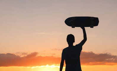 Silhouette of a man holding his longboard skate high against the orange sky after sunset.  Healthier and happier lifestyle through outdoor activities © Marcio
