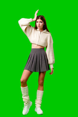 A young woman stands confidently with one hand on her hip and the other touching her head, wearing a fashionable white cropped hoodie and a pleated grey skirt, accompanied by tall white socks. - 788419875