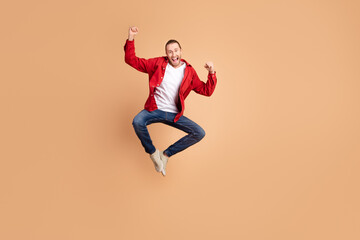 Fototapeta na wymiar Full length portrait of nice young man raise fists jump wear red shirt isolated on beige color background