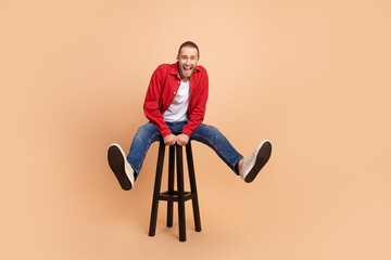 Full length portrait of nice young man laugh sit stool wear red shirt isolated on beige color...