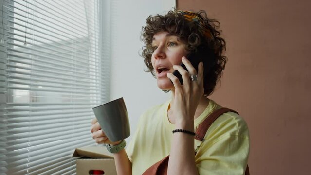 Side chest up footage of smiling Caucasian woman with curly hair and hairband chatting on phone and drinking coffee standing by window in modern apartment and looking away