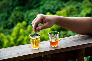A hand is stirring tea in a glass cup, on a wooden bar, green-leaf background