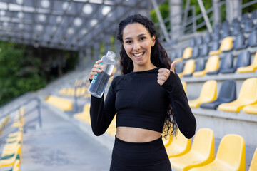 Smiling Hispanic woman with water bottle giving thumbs up after exercise in a stadium. Fitness and...